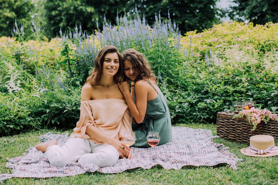 Two young women sit on a plaid blanket outdoors during daytime, surrounded by lush greenery and flowering plants. One woman with medium-length brown hair is smiling and wearing an off-shoulder peach-c