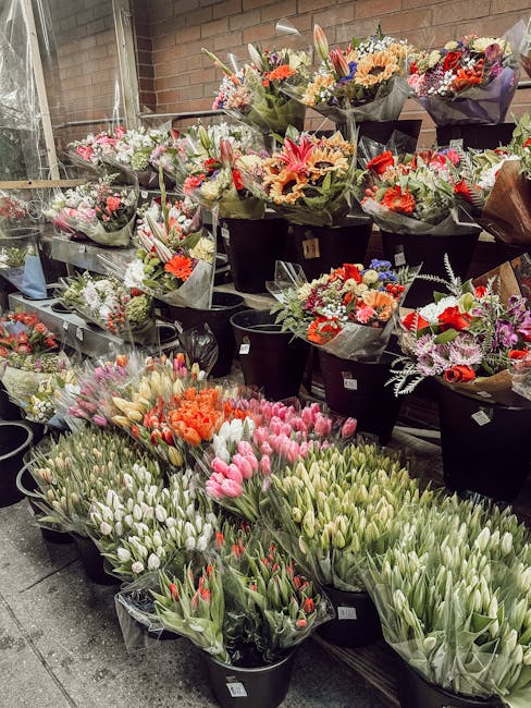A display of various fresh flowers arranged in black buckets at a flower shop, including tulips in shades of pink, white, and red, with some orange flowers. The bouquets feature a mix of blooms such a